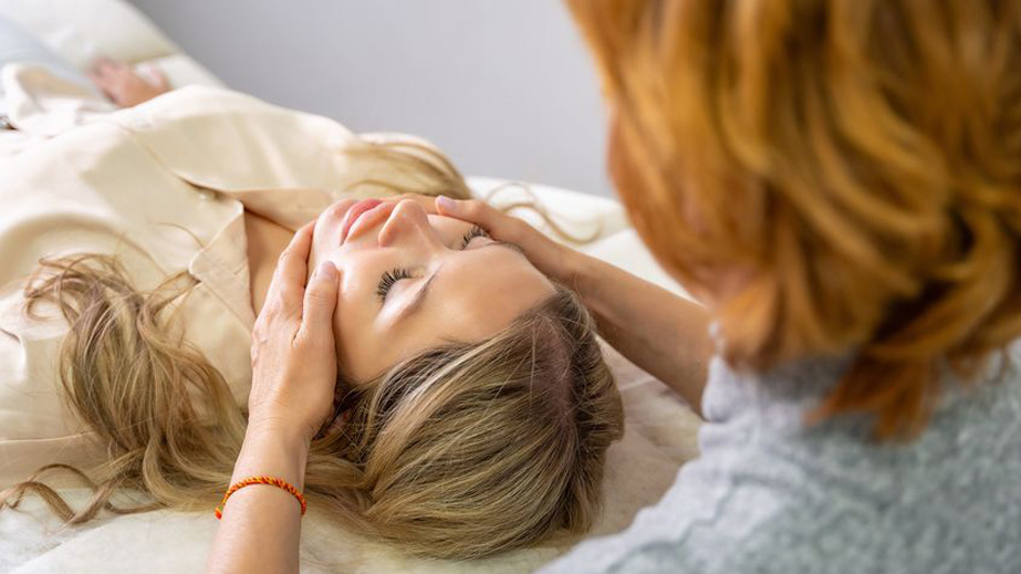 Person lying on a table receiving a facial massage from another individual in a calm setting.