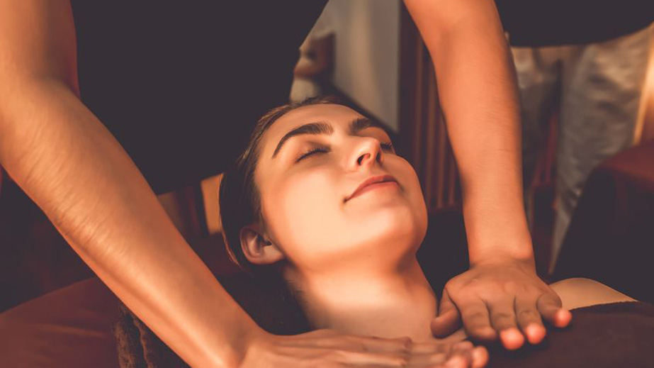 Person with eyes closed receiving a neck and shoulder massage from another individual in a softly lit room.